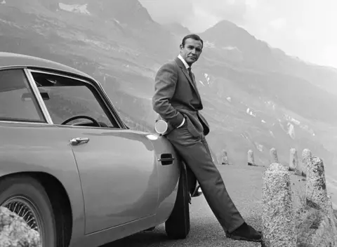 Michael Ochs Archives/Getty Images A black and white still image taken from the James Bond film Goldfinger. Sir Sean Connery stares into the distance as he leans against an Aston Martin DB5 car wearing a suit and tie. Part of a road and a mountain range are visible in the background.