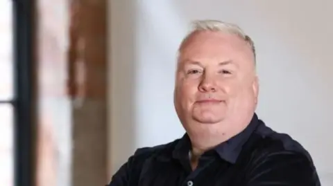 Stephen Nolan filmed in a room with red brick and white walls. He has short grey hair and a black shirt. He is standing with folded arms