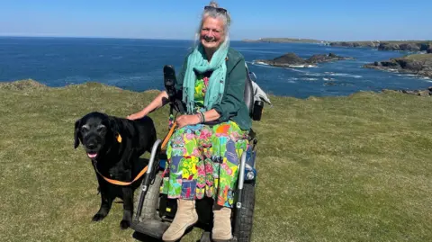 A woman in a wheelchair on a grassy cliff top, overlooking the sea and cliffs with a black dog next to her. She has grey hair and sunglasses on her heard, and is wearing a green jacket, brightly-coloured top and mint green scarf.