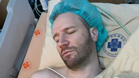 Kevin Smith Kevin Smith is lying in a hospital bed. He has a blue hair net on and his eyes are closed. He has a short beard.