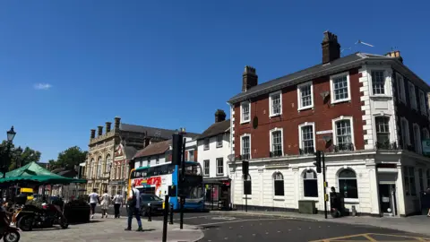 Alex Pope/BBC A town-centre street with a red-brick corner building, a blue-and-white double-decker bus, pedestrians, and market stalls. Historic buildings line the road beneath a bright blue sky