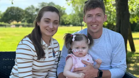 Emily is wearing a cream and light brown stripy top. She is sitting on a bench in the garden with her husband Rory and their daughter, Harriet. Harriet has her hair in pigtails. 