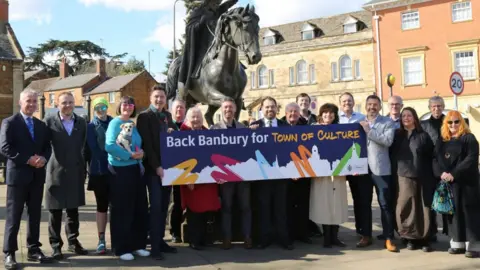 A line of people stood in front of a statue of a lady riding a horse, with some holding a large sign saying 'Back Banbury for Town of Culture'