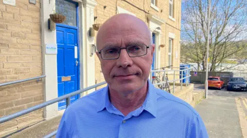 A bald man wearing glasses and a light blue shirt standing outside a building with a blue door.