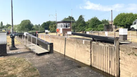 The lock is pictured open and against the side of the river. A brick wall is on the opposite side in the river and a small building with windows stands also on the right side of the water.