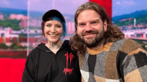 BBC Rox on the left in a black hoodie with red writing on. She has black and blue hair and tattoos on her neck and is wearing a cap. Rich is wearing a checked jumper and has long hair and a beard. They are sat in the BBC South Today studio and are smiling at the camera. 