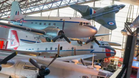 Several historical aircraft are suspended from the ceiling at the museum. They are silver with red paintwork.