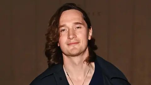 Getty Images Erland Cooper has shoulder-length curly brown hair and smiles at the camera in front of a brown backdrop. He wears a V-beck black shirt and silver necklace.