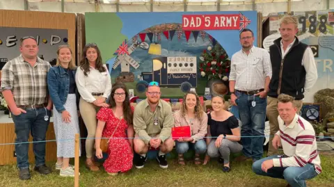 Bea Hodge Ten people in their twenties standing and crouching in front of a Dad's Army display