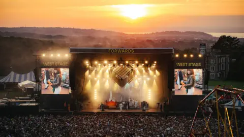 A large crowd watching the West Stage at Forwards. A woman in white jeans and playing a guitar is performing on stage and you can see her on the screens either side of the stage. There a large orange lights shining down on the stage and behind it a beautiful sunset is taking place in similar colours over the horizon.