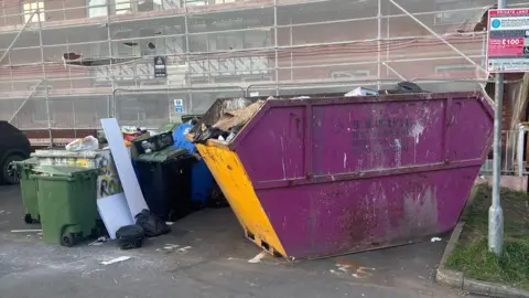 Chris Whittock A purple skip stands outside a building, next to lined-up green bins. The skip is nearly full. The bins are also full and there is debris around. There is scaffolding on the building behind.