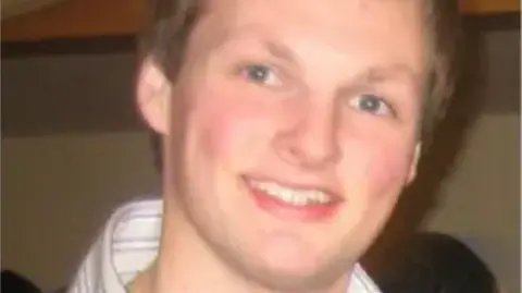 A close-up family photo of the late Shane O’Farrell in which he is smiling at the camera.  The 23-year-old had short, blonde hair and blue eyes.  He was wearing a dark jumper and a blue and white stripped shirt, open at the neck. 