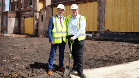 University of Wolverhampton Two men men stand on an area of brown soil with one a spade. On the left, one has a blue suit and brown shoes and the other on the right has dark trousers and a blue shirt on. Both have florescent jackets on and hard hats. Behind them is a boarded-up building and another redundant brick building in the distance.
