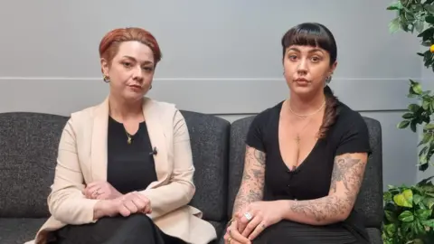 BBC Liz and Catherine McSherry sitting on a sofa - Liz has short red hair - wearing a black top and cream blazer. Catherine has brown hair in a ponytail with a fringe - wearing a black top. 