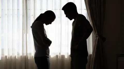 Getty Images The silhouettes of a man and a woman standing in front of a window with sheer curtains drawn. The woman is holding her arms around her waist and looking down, while the man has his hands on his hips and appears to be shouting at her.