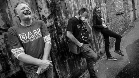 The trio are standing against a wall and laughing together in a black and white photo. They are wearing T-shirts and jeans.