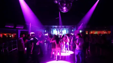 A large group of people on the dance floor of a disco. There is a disco ball on the ceiling and purple spotlights shining on the dance floor.