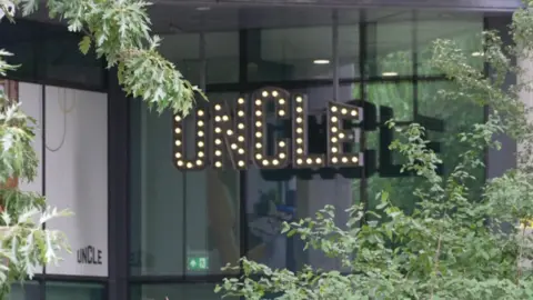 Building entrance featuring a prominent, illuminated marquee-style sign that spells out the word "uncle" in glowing light bulbs, partially framed by green foliage.
