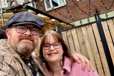 Artisan Tap Scott Evans has his arm around wife Jayne as they stand outside the music venue. He is wearing glasses and a beard. He is wearing a checked coat. Jayne has a pink coat, dark hair and is wearing a necklace. They are both smiling. 