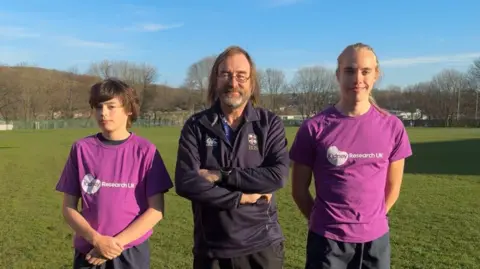 Kate Stanley Two teenage boys wearing a purple t-shirt with a white logos and a man standing in the middle of them with long, brown hair and wearing a blue sports top. 