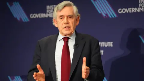 Getty Images Former British Prime Minister Gordon Brown speaks at the Institute for Government on March 11, 2024 in London, England on the urgent challenges facing the UK government. He is wearing a navy suit jacket, light-blue shirt and red tie. He has grey hair. 