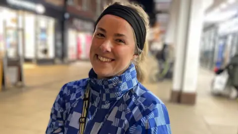 Laura-May Land wears a black hairband, a blue patterned jacket and has a strap across her shoulder. She is smiling directly at the camera and has her head tilted to the side. She is standing in Cambridge city centre.