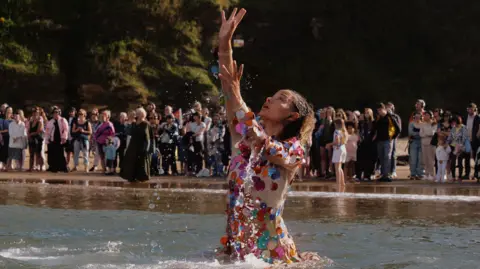 Luke Waddington A woman performs a dance in the sea in front of a crowd of people gathered on the beach.