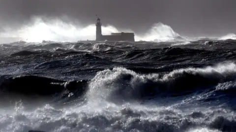 PA Media Giant dark blue waves hit the lighthouse wall at Whitehaven when a storm hit the area in 2014. It is a very dark and stormy day.