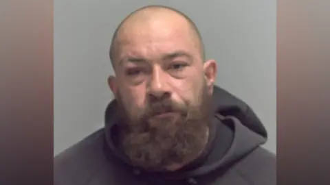 A custody image of a man with a shaved head and a thick beard. He is wearing a grey hoodie.