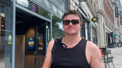 Sofía Luis-Hobbs/BBC A man wearing a black tank top and black sunglasses smiles for the camera on a paved high street.