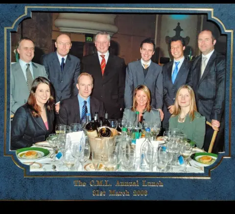 Picture of REWS in a group smiling on camera. Reeves is sitting next to a man, and two other women are sitting on a table, with bottles and glasses of wine. Six people are standing behind the group sitting in a semi circle.