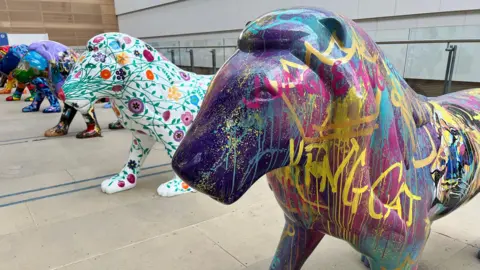 A row of the painted lions inside a large inside space. They are all brightly coloured, and the one closest to the camera is purple and covered with a graffiti pattern. The one next to it is white, covered with green leaves and multicoloured flowers.