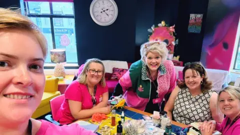 Support 2gether Group leader Ciara is holding the camera. She has short blonde hair and is taking a selfie with four women in the background. Four of the women are wearing pink tops with the charity logo. There are crafts on the table.