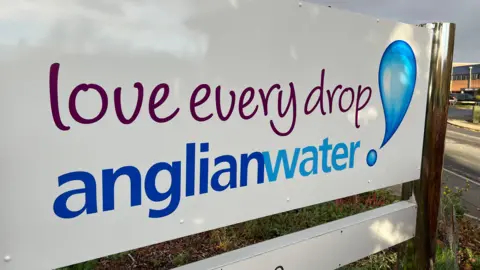 A white sign with purple and blue writing. It reads 'Anglian Water, Love every drop'. There is a exclamation mark at the end which had been designed to look like two droplets of water. The sign is outside the companies HQ