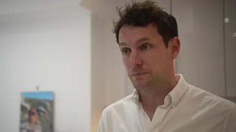 BBC A man with brown hair in a white shirt holds a serious expression. He appears to be in a living room.
