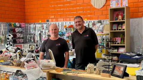 Two men - Colin and Francis - stand smiling in the craft shop where they work. They are surrounded by an assortment of handmade goods.