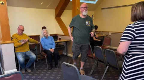 Simon Thake A group of men and women in everyday clothes rehearse a play in an attic space with wooden beams and low ceilings. An elderly woman in a blue sweater sits, watching thoughtfully