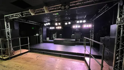 Tom Jackson/BBC A stage is situated in the corner of a room. There is stage lighting on the ceiling and walls surrounding the stage. On both sides, there are black boxes and a silver railing.