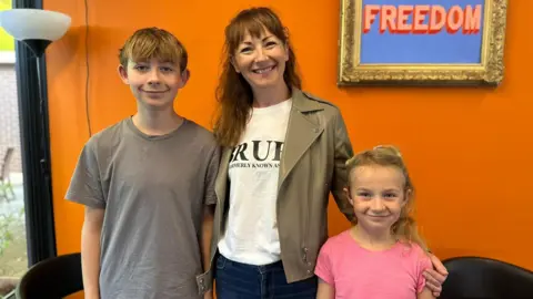 Coletta Todd with long ginger hair and a fringe standing between her 14-year-old son henry who is wearing a grey t shirt and her sweven-year-old daughter Mary who is wearing a pink t-shirt. They are standing in front of a bright roange wall with a lamp to one side and gold frame on the wall with the words Freedom in typographical art