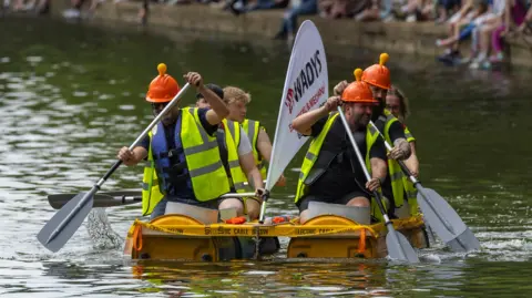 Bedford Borough Council Six people on a raft, on the water, all wearing high-vis jackets, wearing hard hats and holding paddles. People can be seen with their legs over a wall behind them.