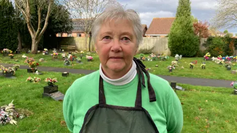 Liz, a woman with short grey hair, stands in a graveyard with green grass and headstones around her. She wears a green jumper with a darker green apron on top.