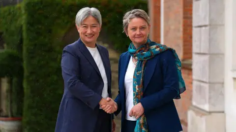 Getty Images Two women are shaking hands and looking at the camera. Both have navy blue suits on and white t-shirts. Both have short grey hair. The woman on the right has a bright blue scarf. Behind them is a red brick building