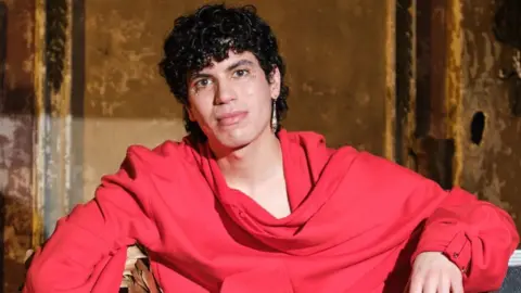 Jacob Alon, wearing gold feathered trousers and an oversized red sweater, poses on flight cases backstage at the BBC's Jools Holland show