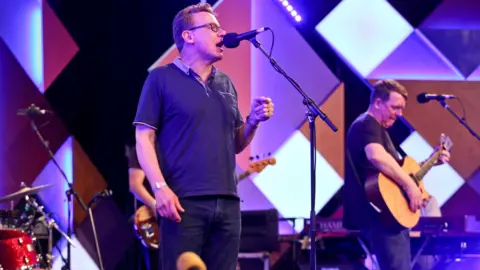 Tricia Yourkevich/BBC The Proclaimers on stage with purple and blue lighting and a background of diamonds and triangles. The singer and guitarist each stand behind a microphone.