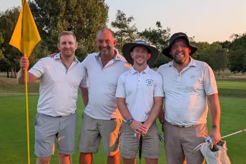 Jonathon Wallace, Mike Ashwin, Ben Read, and Ben Smith pictured at Chipping Sodbury Golf Club, wearing white polo shirts and khaki shorts, standing on the green after completing their16-hour golf challenge with rolling fairways and trees behind them.