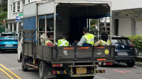 BBC/Gavin Butler Um grupo de trabalhadores migrantes sentados no fundo de um caminhão em Cingapura