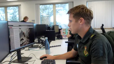 Cameron in the control room at Yorkshire Ambulance Service, where he works as a a logistics team leader, often training before and after work. He's in profile, wearing a dark green uniform, and working at a computer. Behind him are windows