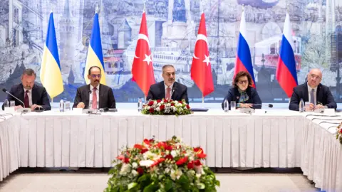 Delegates from Turkey, Ukraine and Russia sit at a table with flags behind and flower arrangements in front of them