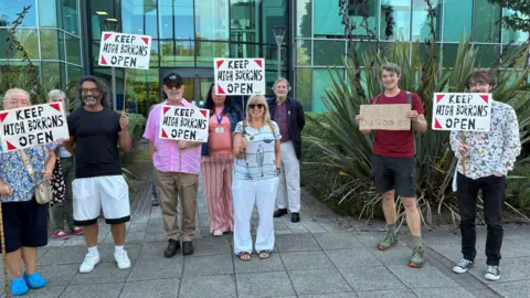A group of nine men and women gathered outside the headquarters of North Tyneside Council. They are holding small placards reading "Keep High Borrans open". The building behind them has glass frontage and there are spiky plants in front.