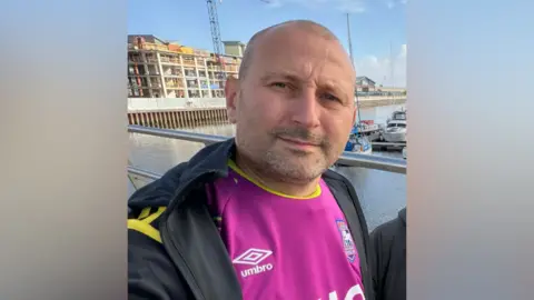 Supplied Paul Munnings wearing a pink Ipswich Town football top under a black and yellow zip-up top. He is smiling into the camera while standing in front of what looks like a marina with boats.